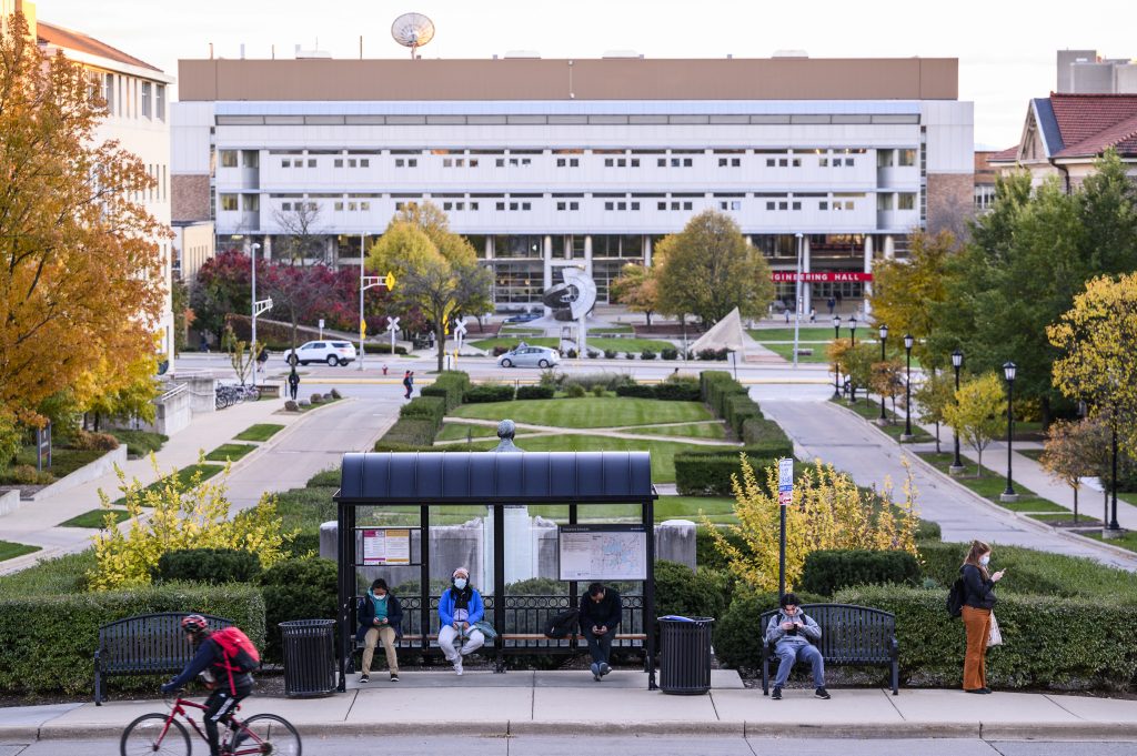 People wait for a Madison Metro bus at a transportation stop along Linden Drive and Henry Mall at the University of Wisconsin–Madison during an autumn sunset on Nov. 2, 2021. On the horizon are vehicles traveling along University Avenue plus Engineering Mall and Engineering Hall. (Photo by Jeff Miller / UW–Madison)